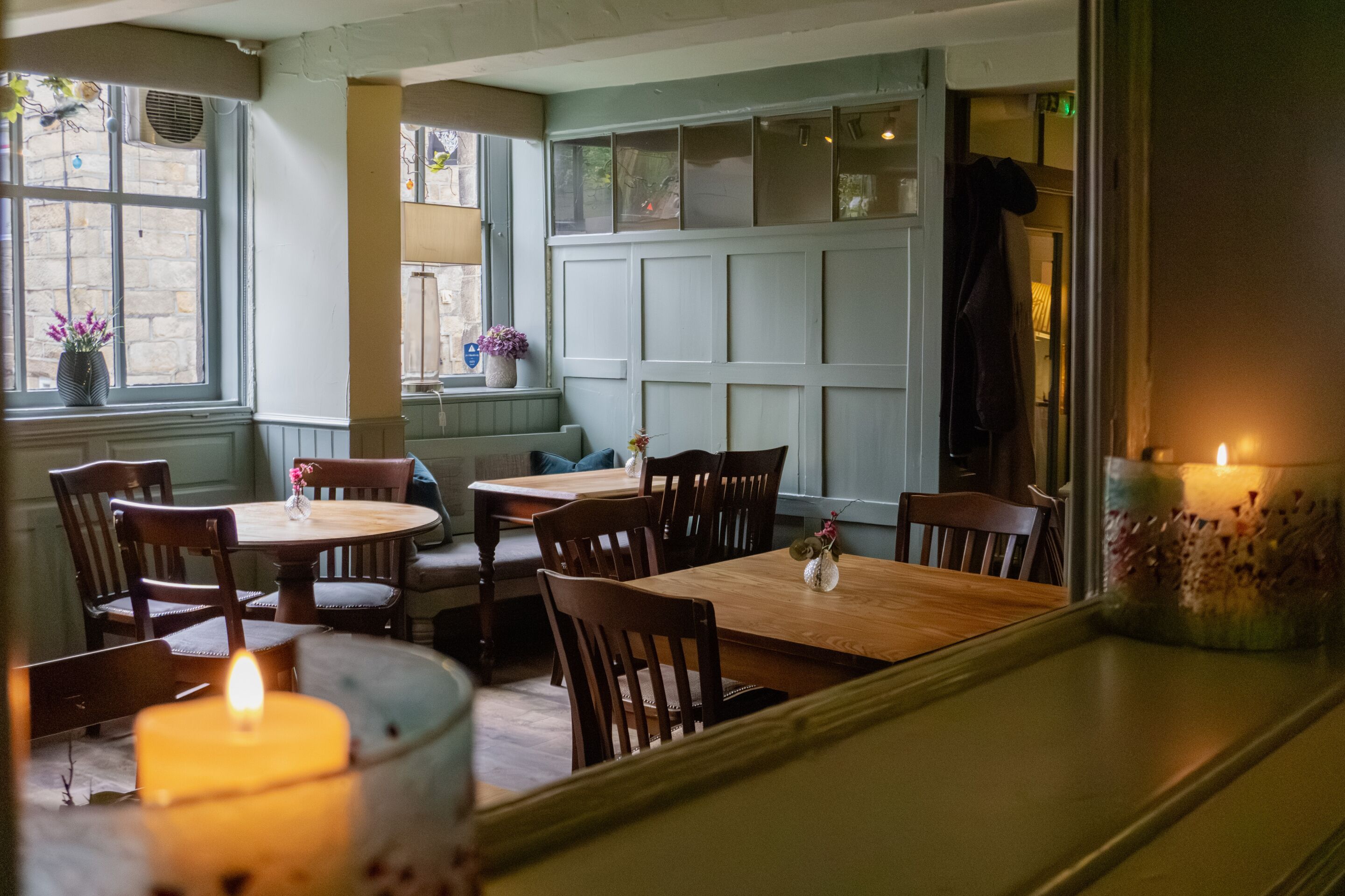 A cozy dining area with wooden tables and chairs, a large window with a view, and a candle on the table.