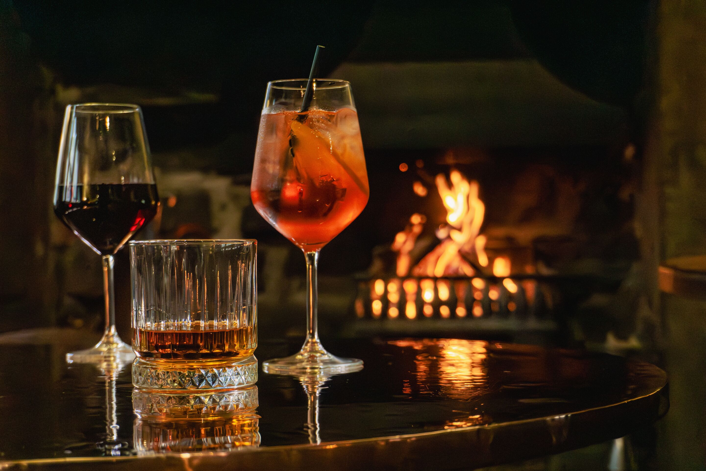 Three glasses of beverages on a reflective surface with a fire in the background.