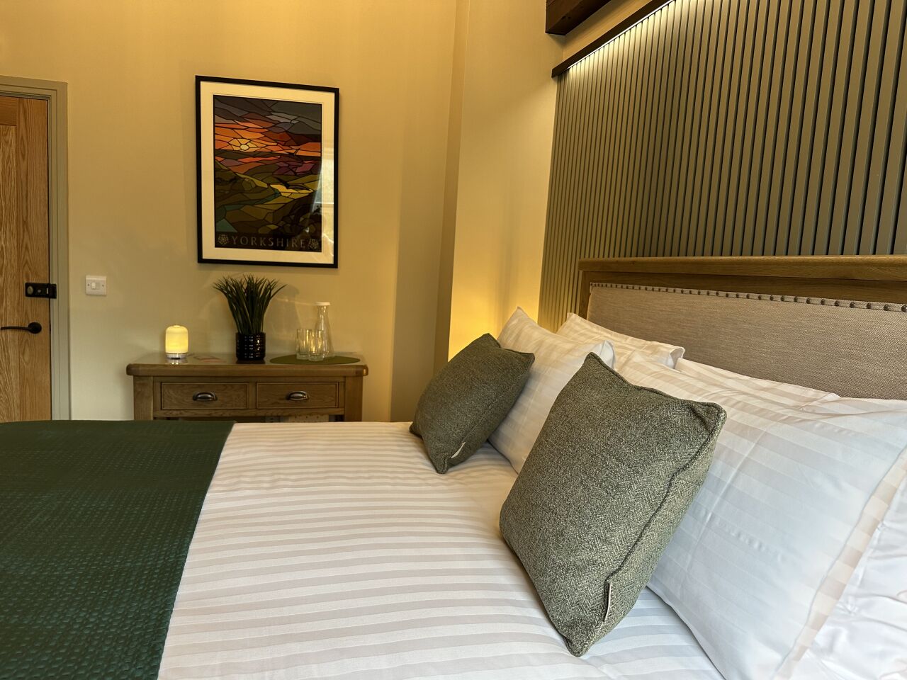 A neatly made bed with striped bedding, a headboard with vertical slats, a framed picture on the wall, and a bedside table with a lamp and decorative items.