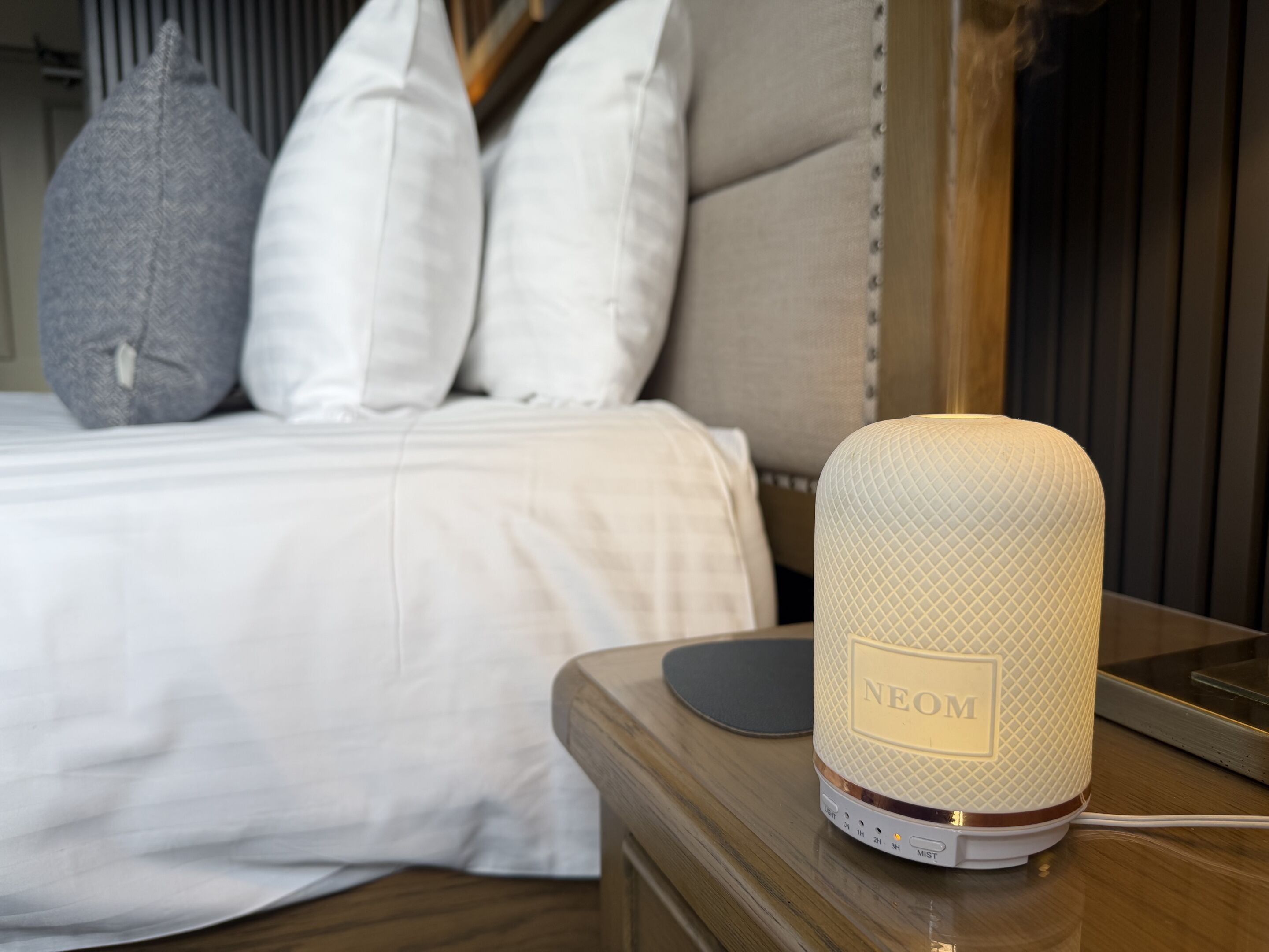 A bed with white bedding and pillows, a bedside table with a white lamp labeled "NEOM," and a wooden nightstand.