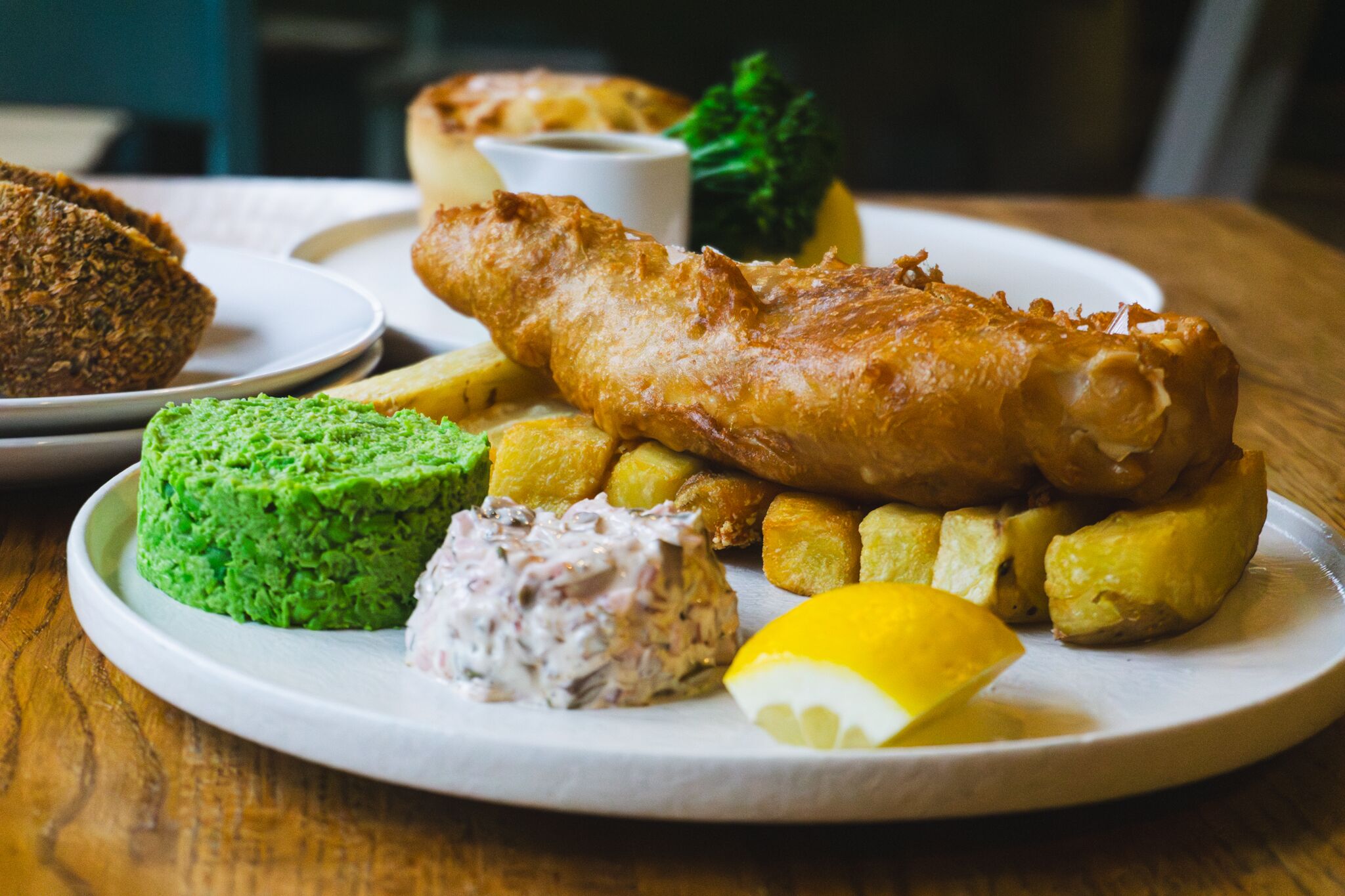 Um prato de peixe com batatas fritas, acompanhado de ervilhas amassadas, molho tártaro e uma fatia de limão.
