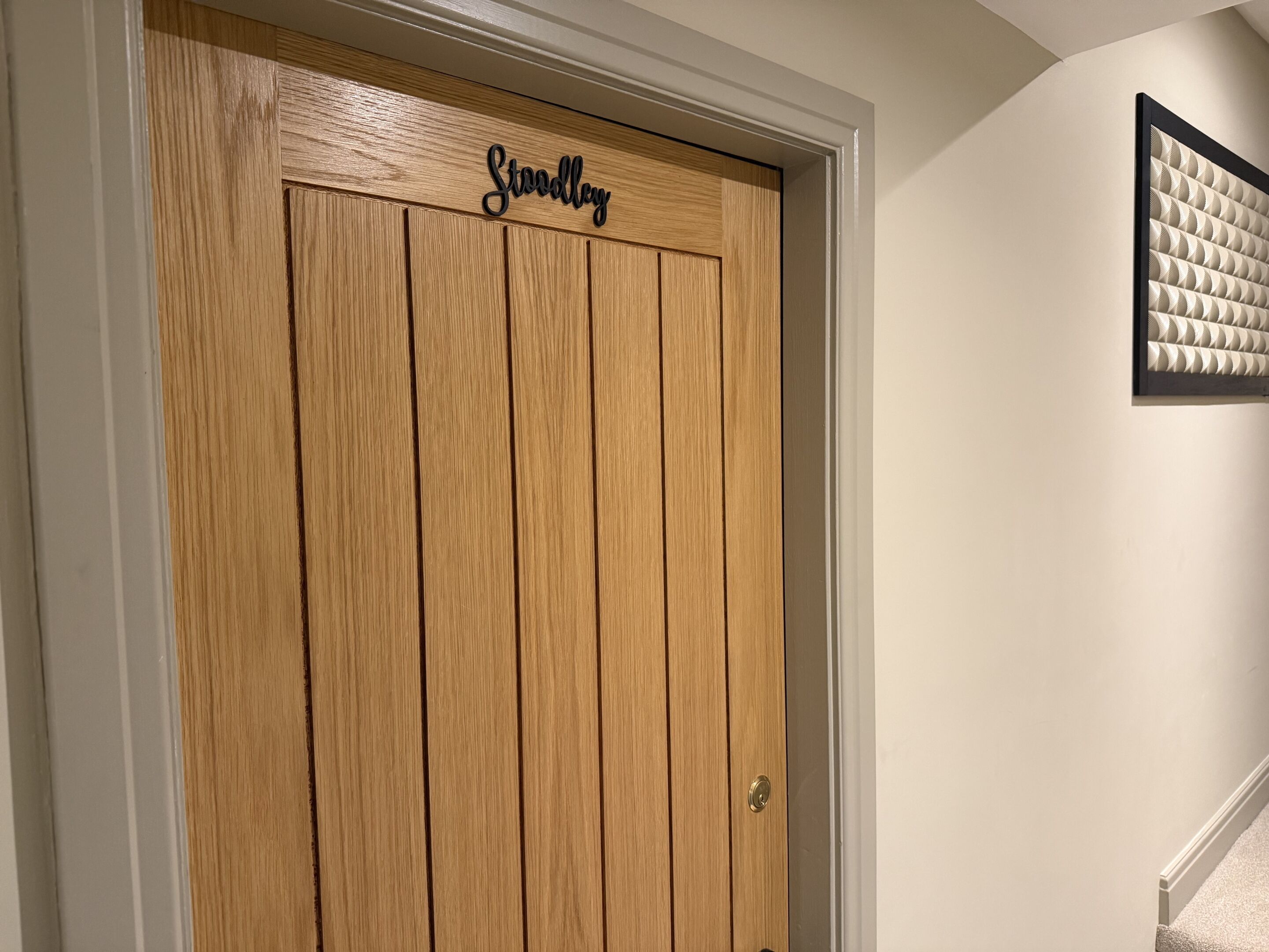 A wooden door with the name 'Stoddley' carved on it, a white wall, and a framed artwork above the door.