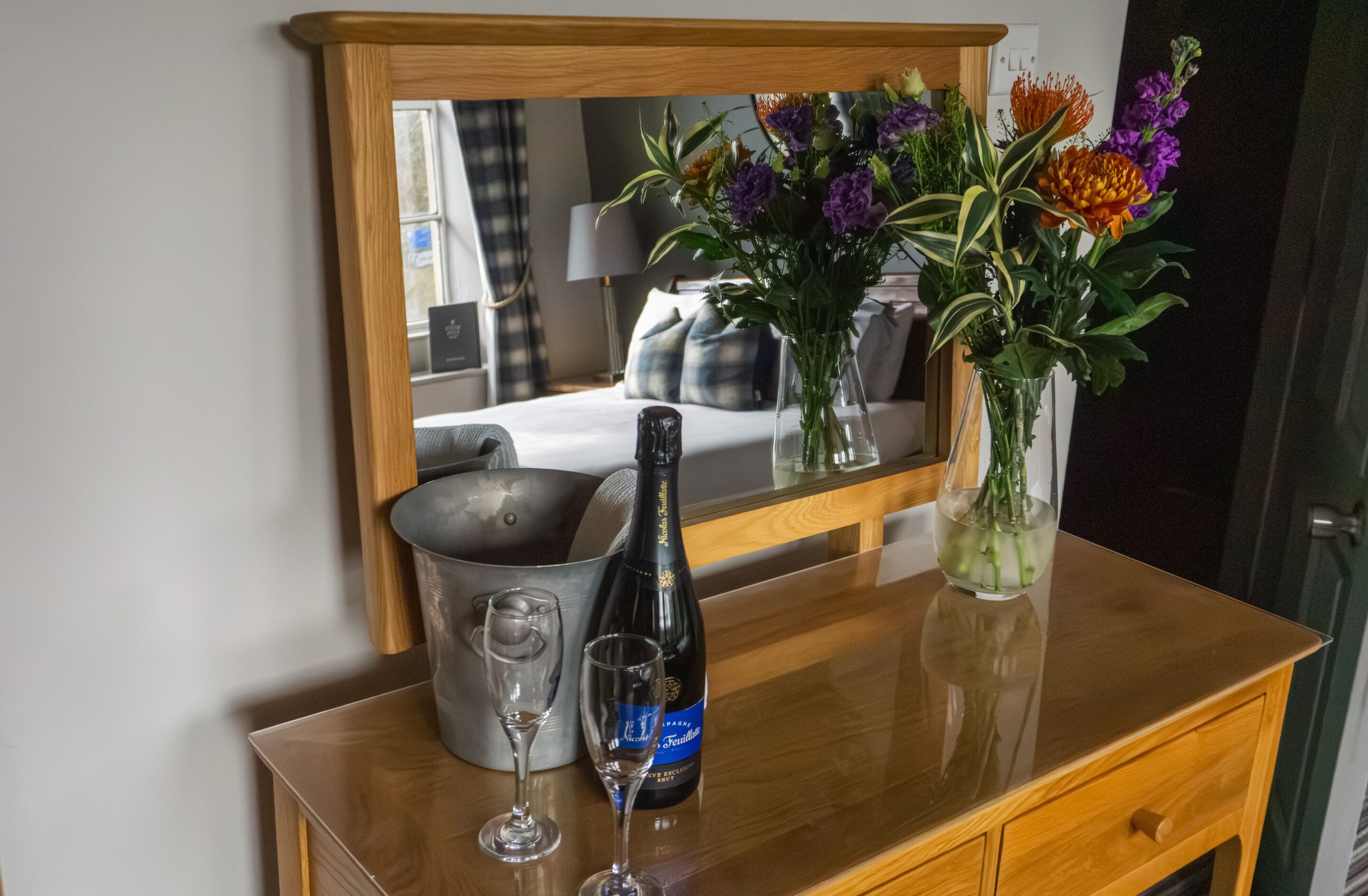 A wooden dresser with a mirror, a bottle of champagne, two wine glasses, and a vase with flowers.