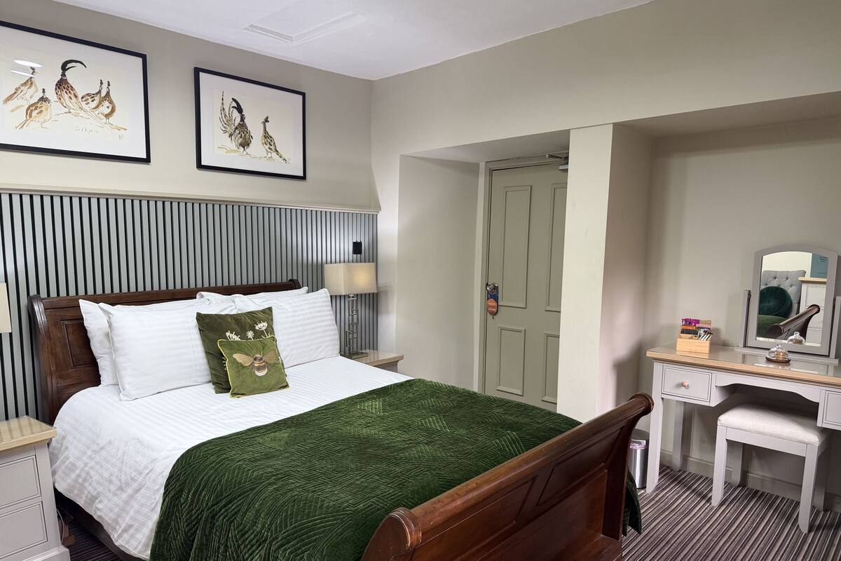 A hotel room with a bed, a desk, a chair, a lamp, and two framed pictures on the wall.