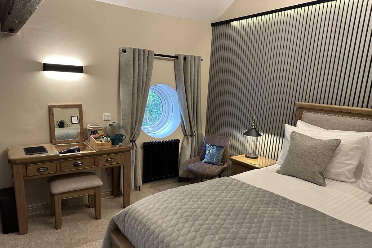 A bedroom with a bed, a desk, a chair, a nightstand, a lamp, and a window with curtains.