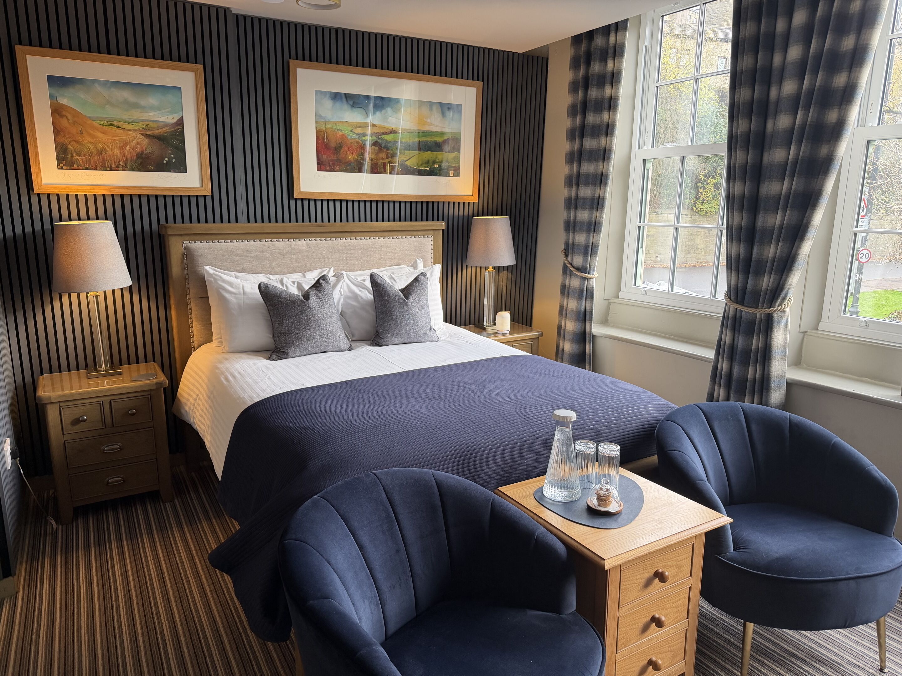 Une chambre d’hôtel avec un grand lit, deux lampes de chevet, une table de nuit, une petite table en bois avec une lampe, deux chaises et deux tableaux représentant des paysages, accrochés au mur.