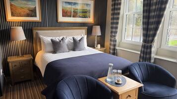 Une chambre d’hôtel avec un grand lit, deux lampes de chevet, une table de nuit, une petite table en bois avec une lampe, deux chaises et deux tableaux représentant des paysages, accrochés au mur.