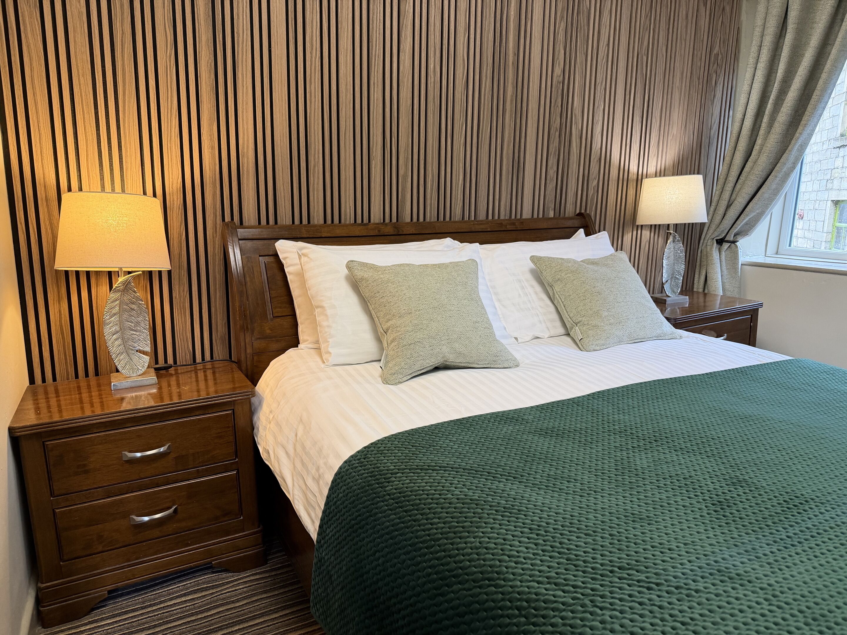 A neatly made bed with white linens and two pillows, a wooden nightstand with a lamp, and a green textured throw blanket.