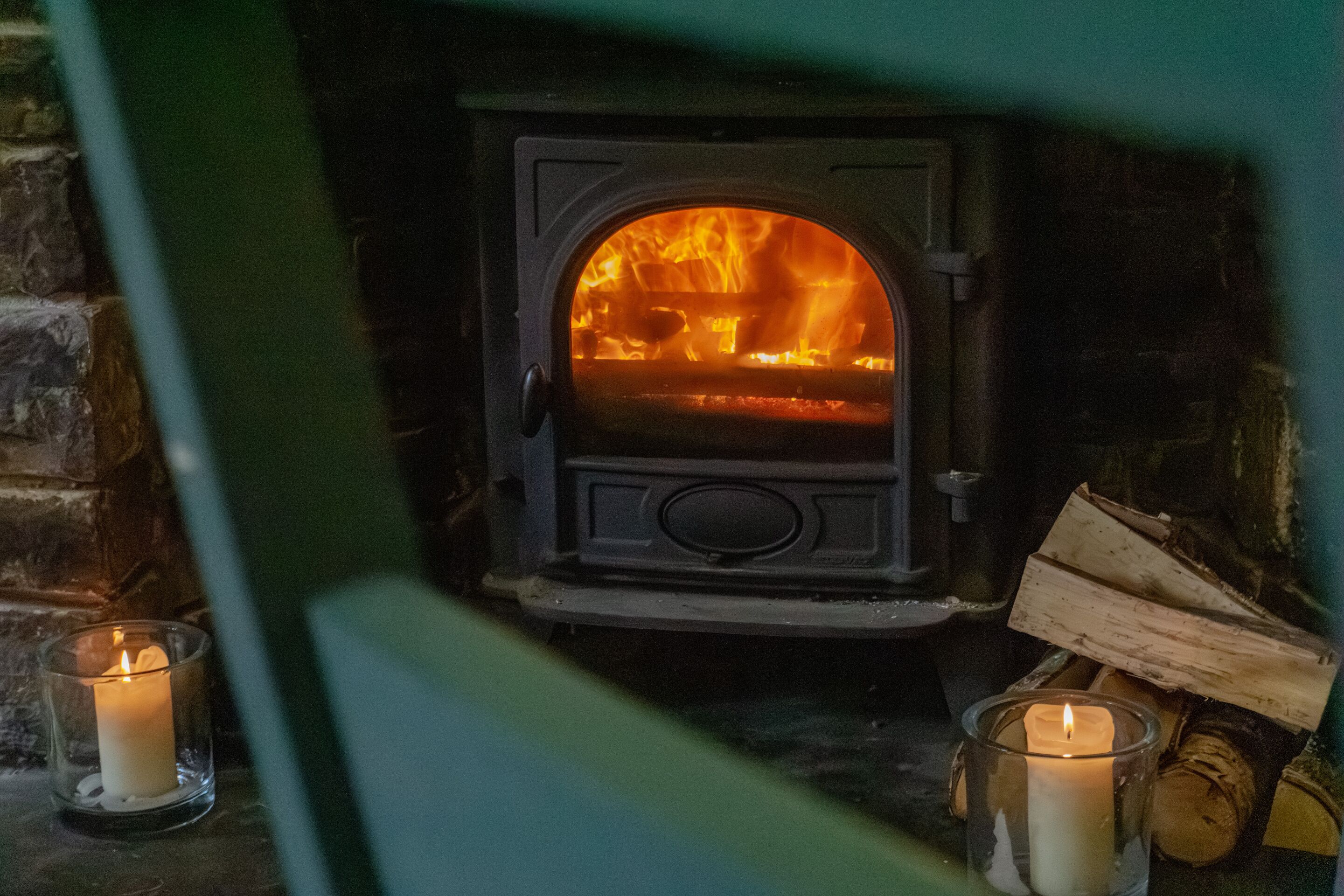 A wood-burning stove with visible flames, two lit candles, and stacked firewood.