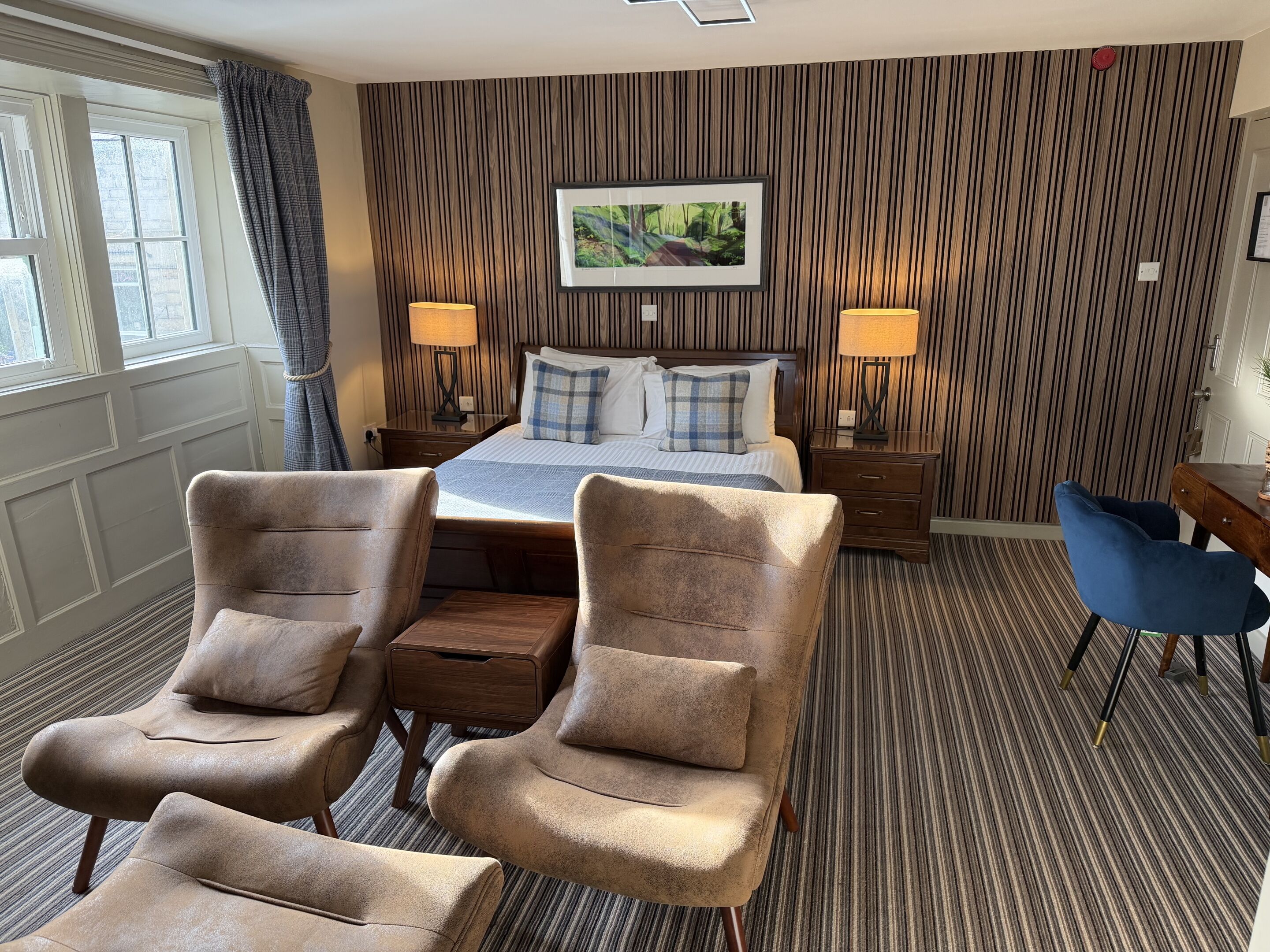 Habitación de hotel con cama, dos mesitas de noche, dos lámparas, un escritorio, dos sillones y una ventana con cortinas.