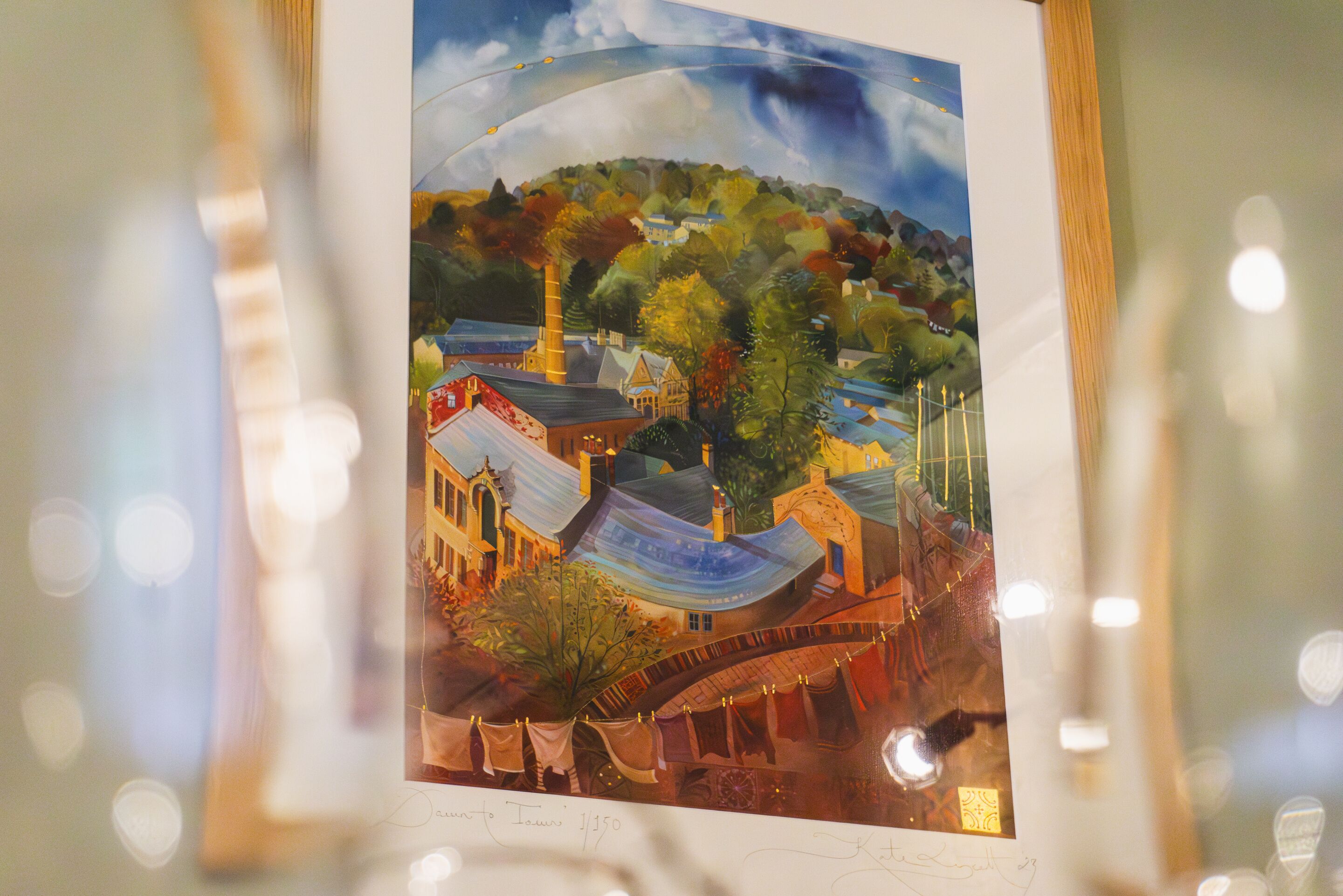 A framed painting of a village scene with traditional buildings, a bridge, and a hill in the background.