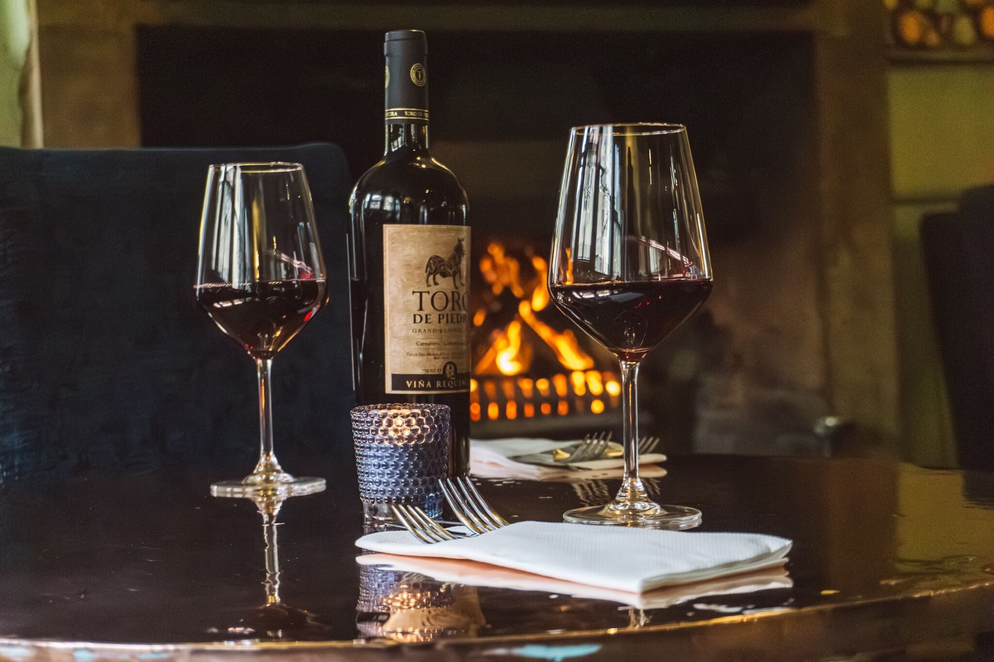 A bottle of wine, two wine glasses, and a glass of water on a table with a fireplace in the background.
