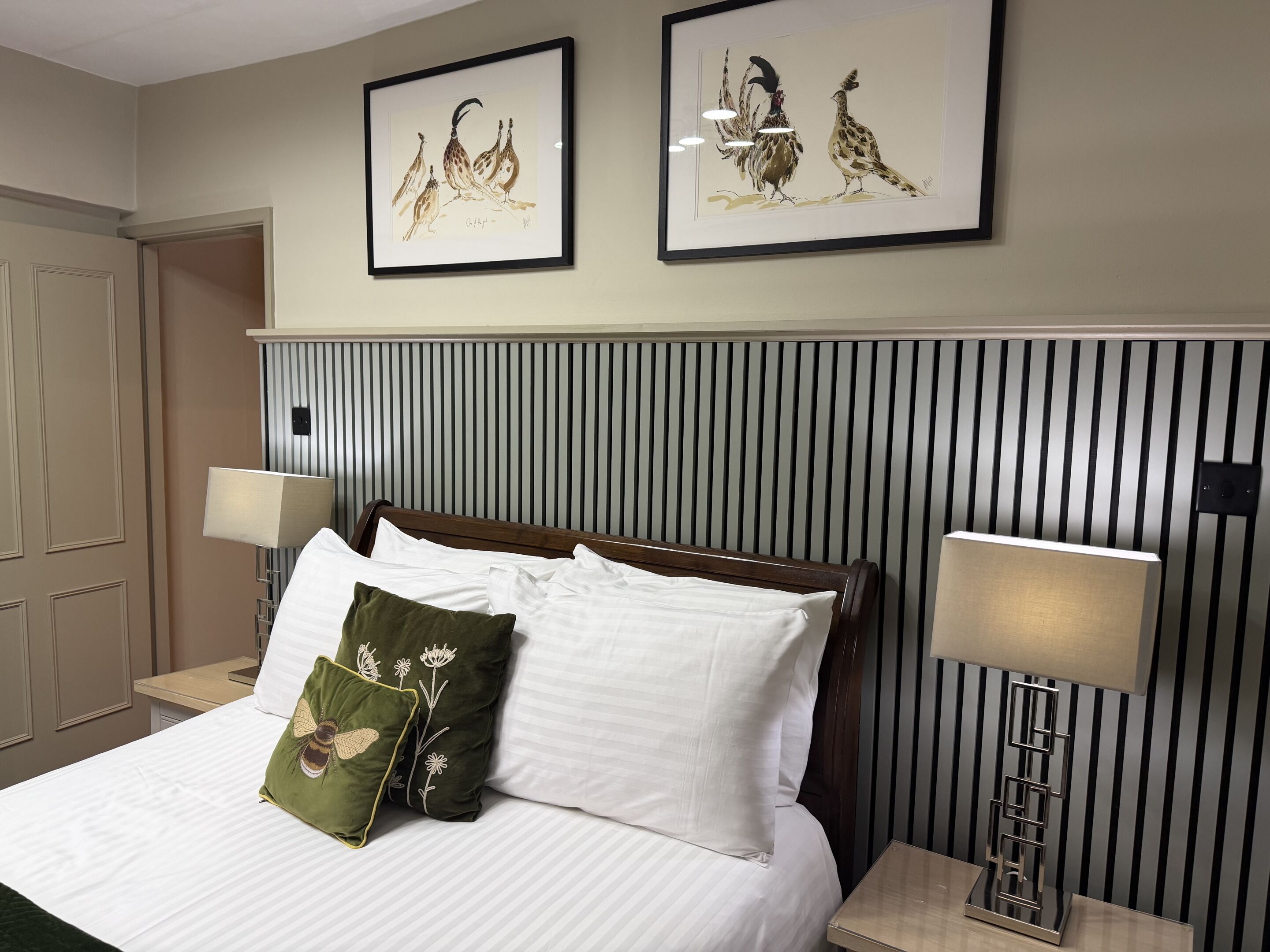 Ein Hotelzimmer mit einem Bett, zwei gerahmten Kunstwerken, einem Nachttisch mit Lampe und einer Wandleuchte.