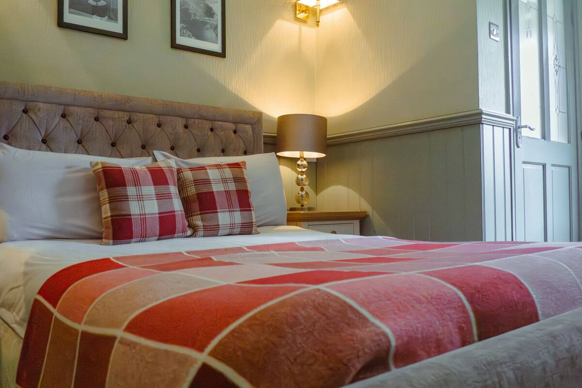 A neatly made bed with a red and white checkered bedspread, a plaid pillow, and a wall-mounted lamp.