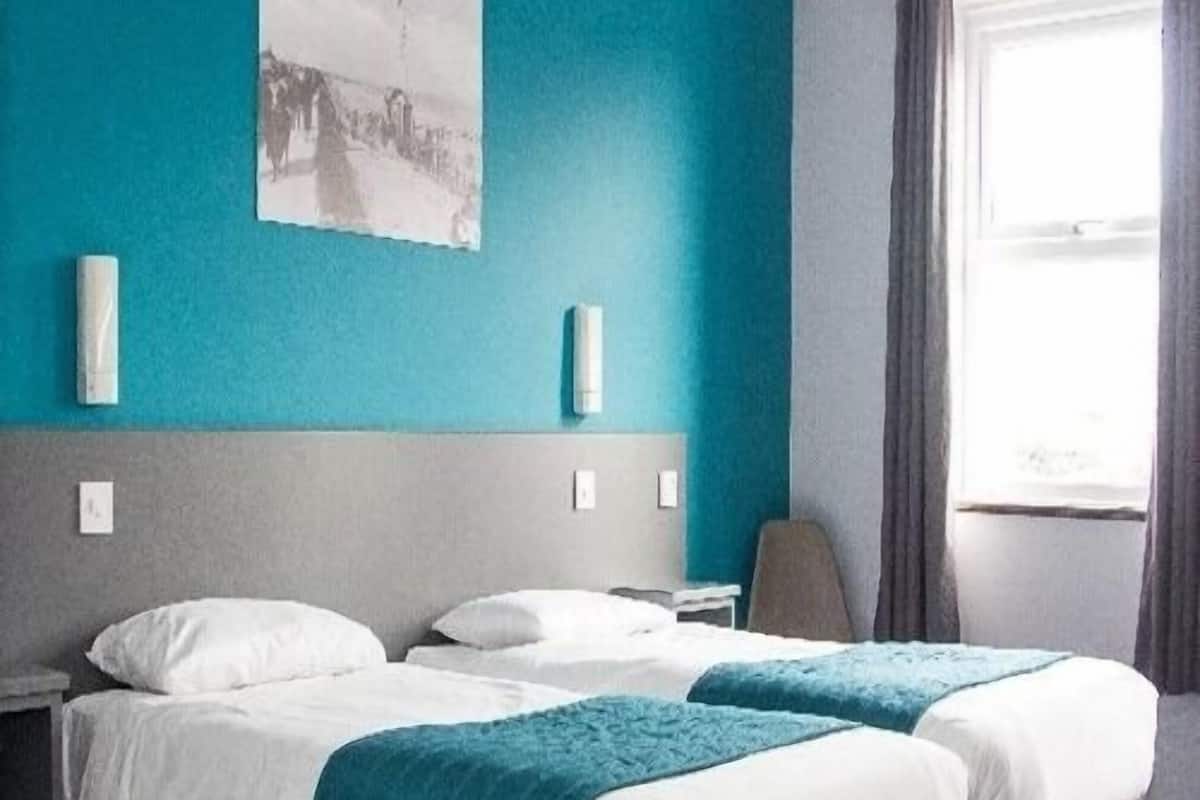 A hotel room with two beds, a grey headboard, a teal accent wall, and a window with curtains.