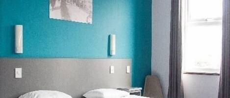 Ein Hotelzimmer mit zwei Betten, einem grauen Kopfteil, einer türkisfarbenen Akzentwand und einem Fenster mit Vorhängen.