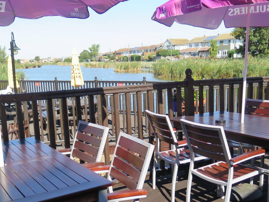 An outdoor dining area with wooden tables and chairs, purple umbrellas, and a view of a body of water and houses in the background.
