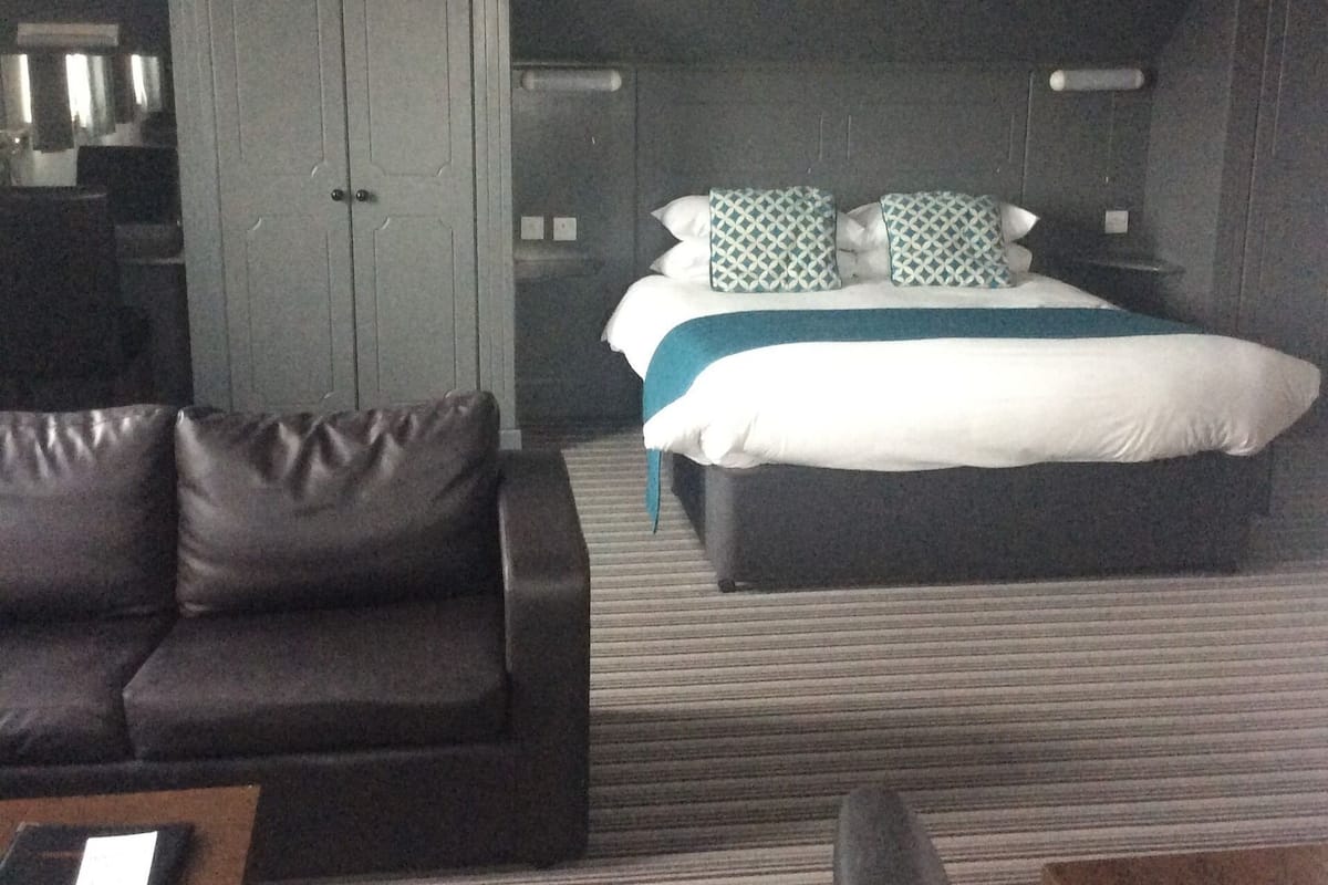 A hotel room with a dark brown leather sofa, a bed with a white and teal bedspread, and a grey wardrobe.