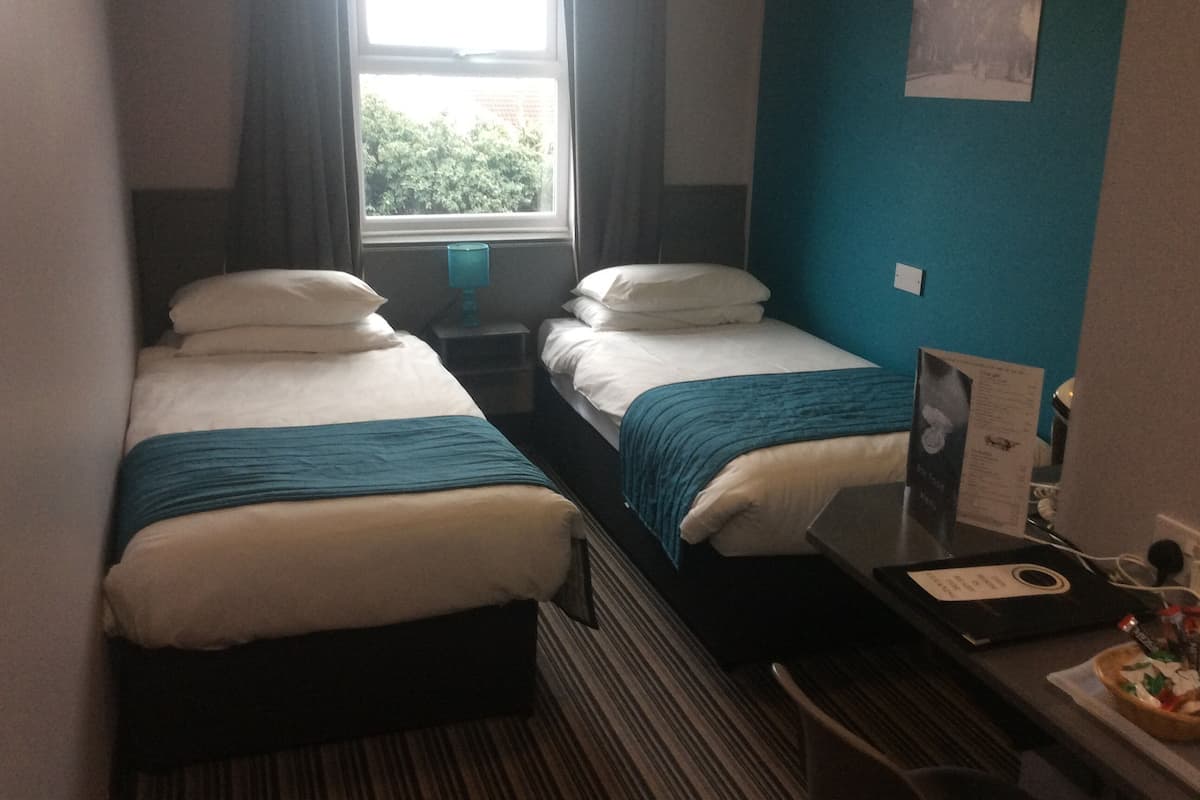 A small hotel room with two single beds, a window with curtains, a teal wall, and a small table with a menu.