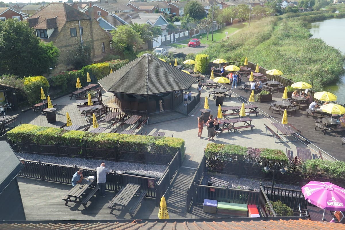 A riverside outdoor seating area with tables and umbrellas, surrounded by residential buildings.