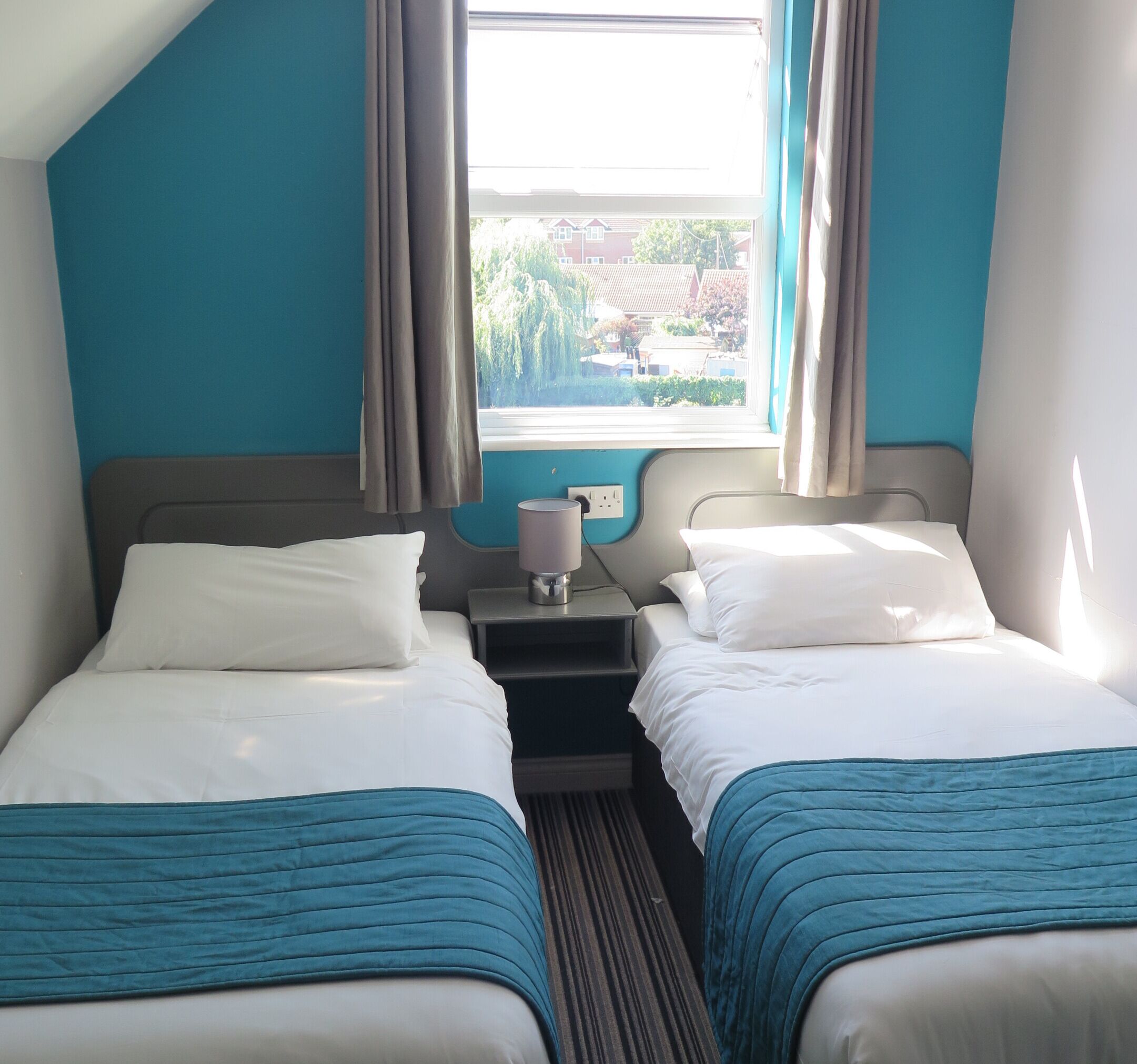 A hotel room with two beds, a window with curtains, a bedside table with a lamp, and a view of the outside.