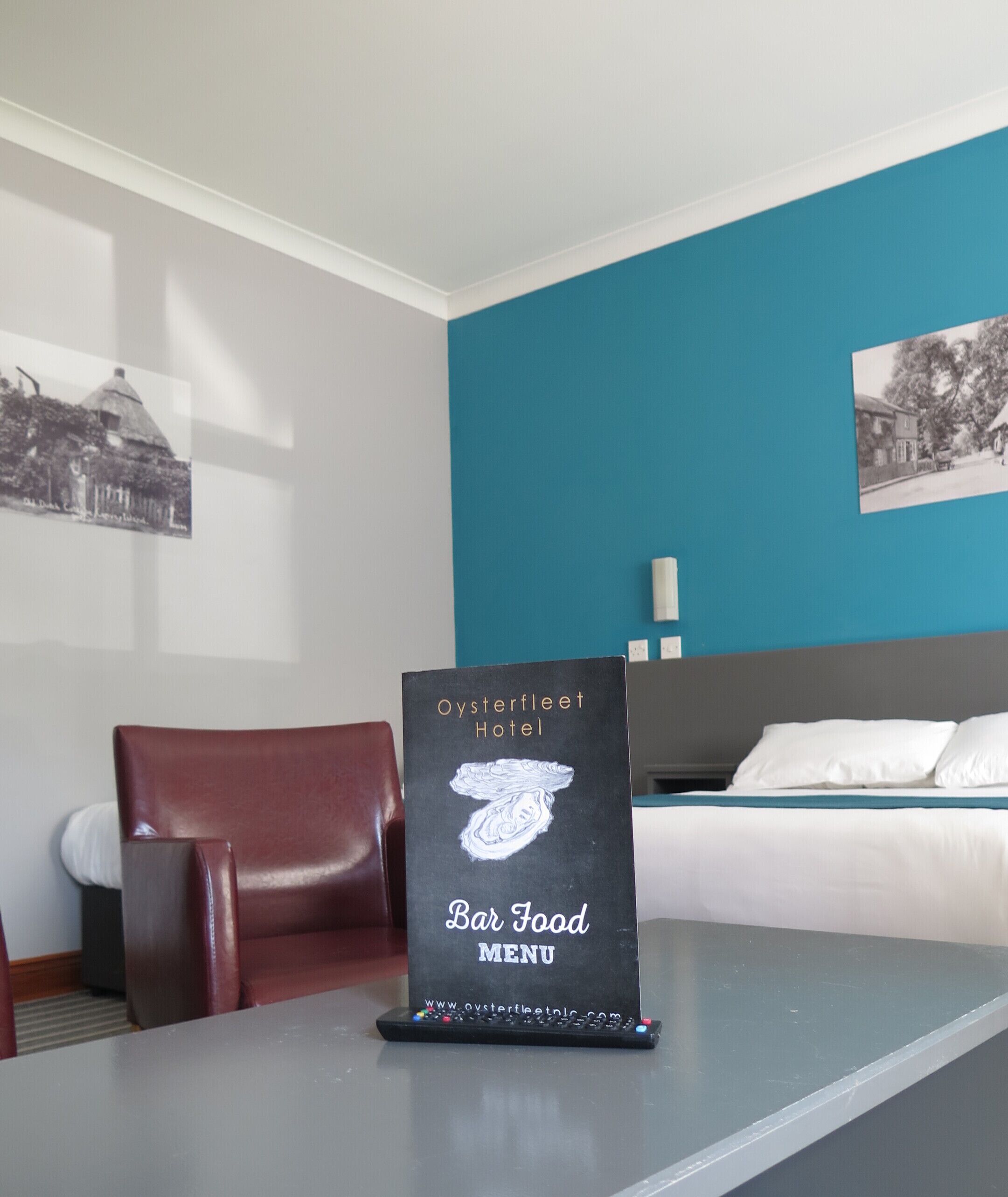 A hotel room with a bar food menu stand, a bed, a red chair, and a wall with a black and white photo.