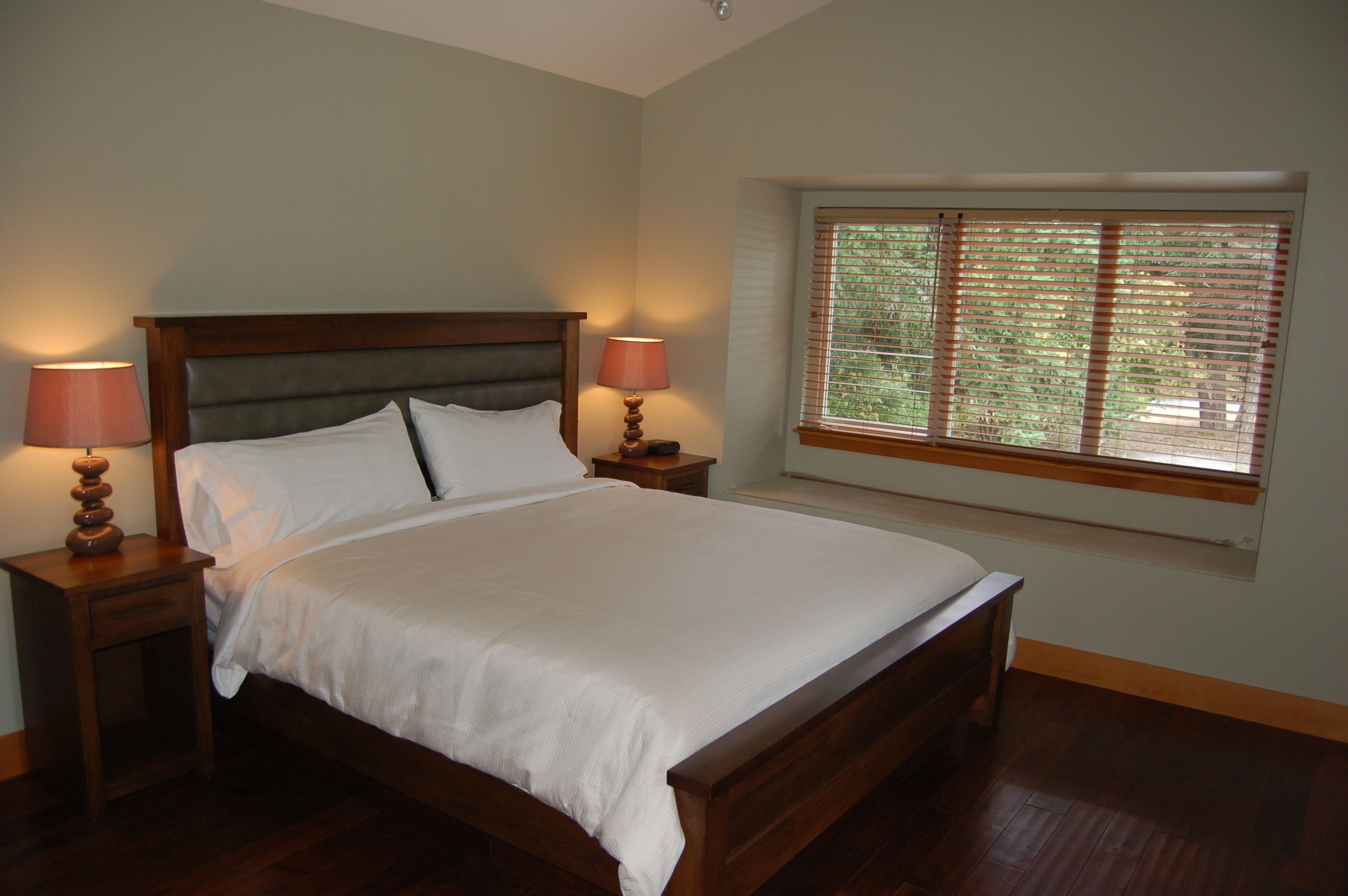 A neatly made bed with white linens, two bedside lamps, and a window with blinds.