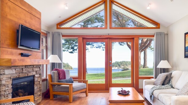 A living room with a stone fireplace, a television mounted on the wall, a sofa, a coffee table, and large windows offering a view of the ocean.