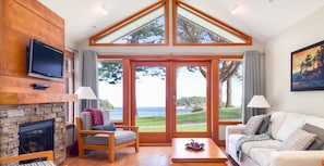 A living room with a stone fireplace, a television mounted on the wall, a sofa, a coffee table, and large windows offering a view of the ocean.