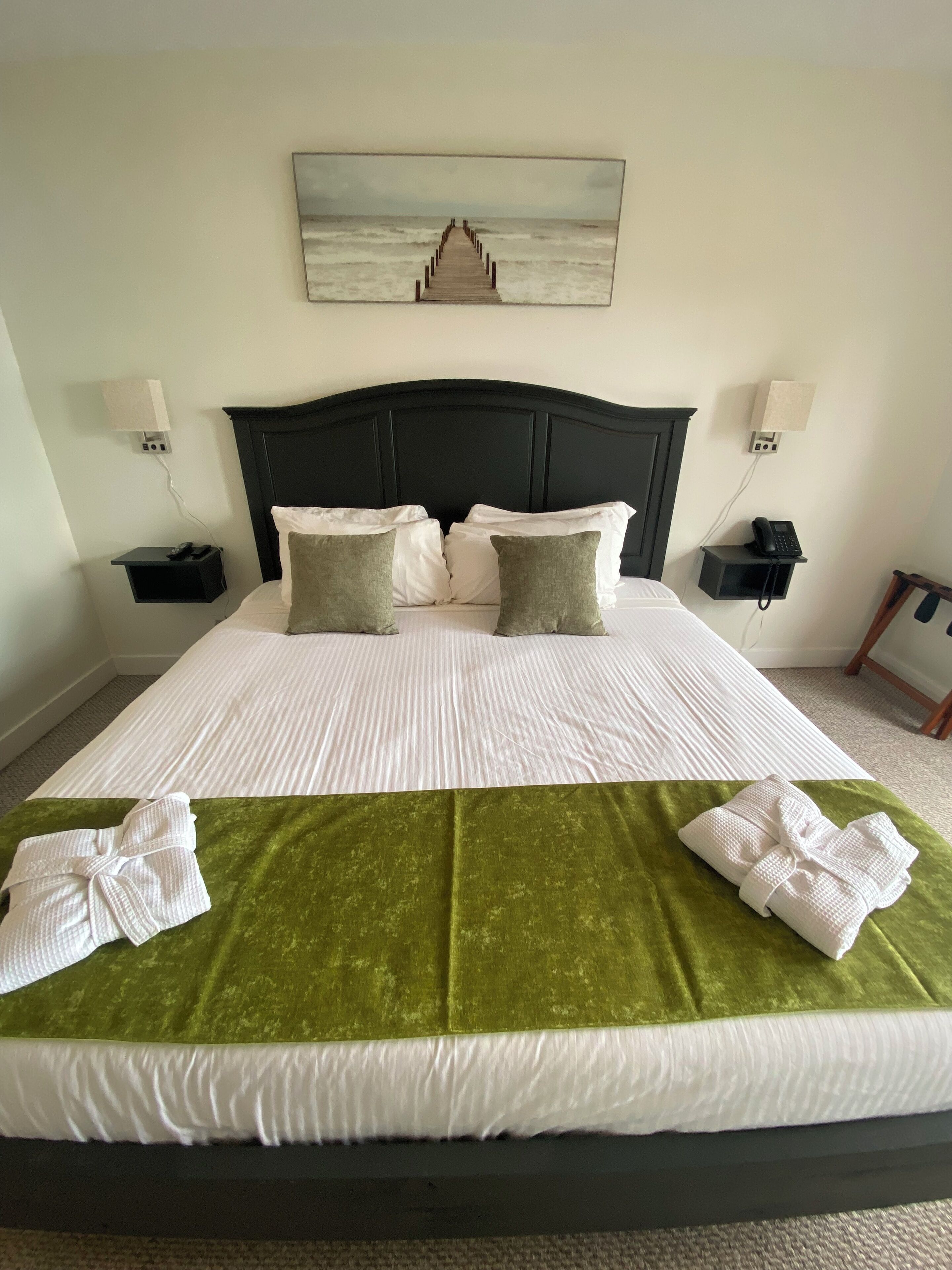 A neatly made bed with a green blanket and two white towels, a dark headboard, and a painting of a pier on the wall.
