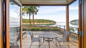A covered outdoor patio with a round table and four chairs, overlooking a lake and a forested area.