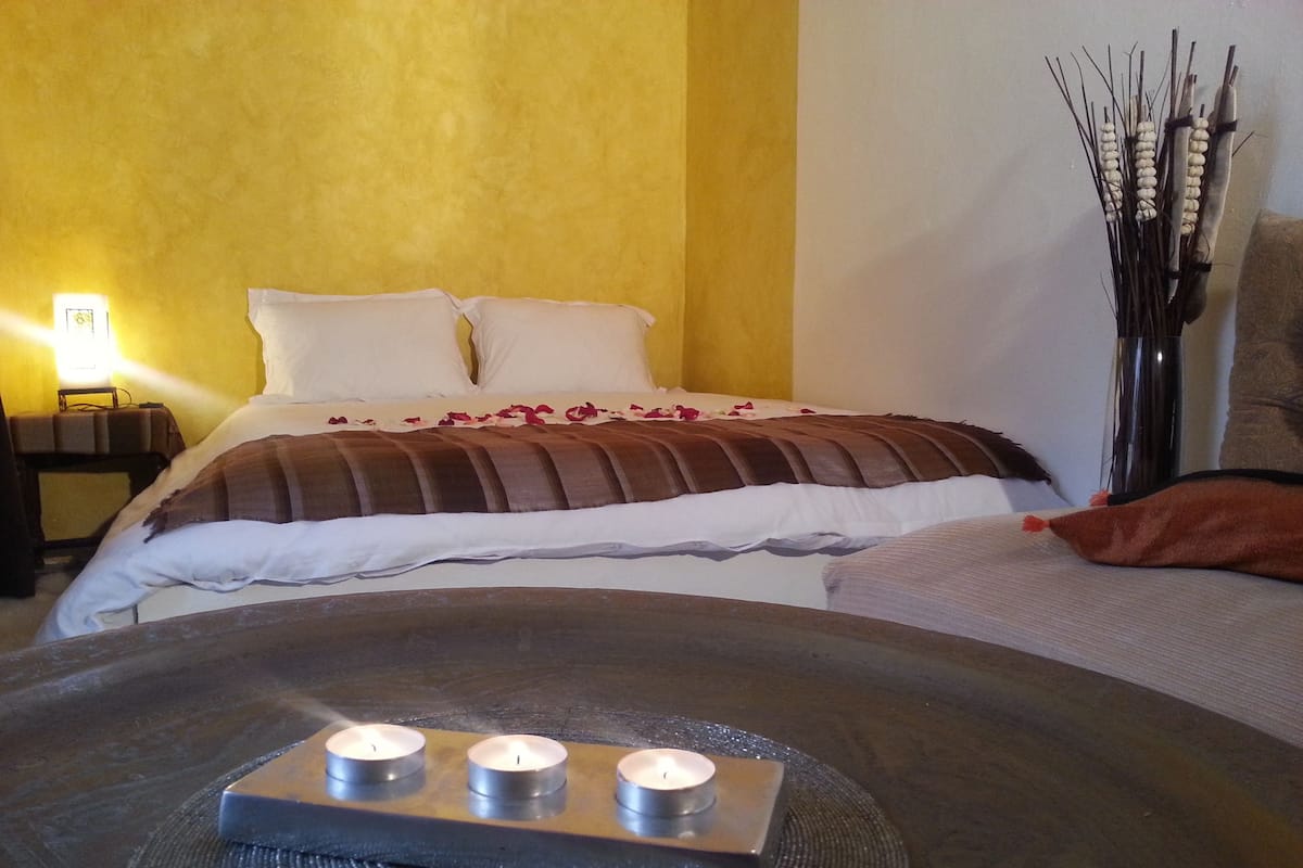 A neatly made bed with white and brown bedding, a bedside table with a lamp, and a decorative vase with branches on the right side.