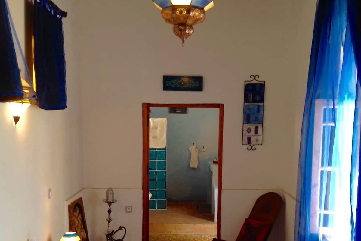 A bedroom with a bed, a nightstand, a lamp, a chair, a wardrobe, a mirror, and a door leading to another room.