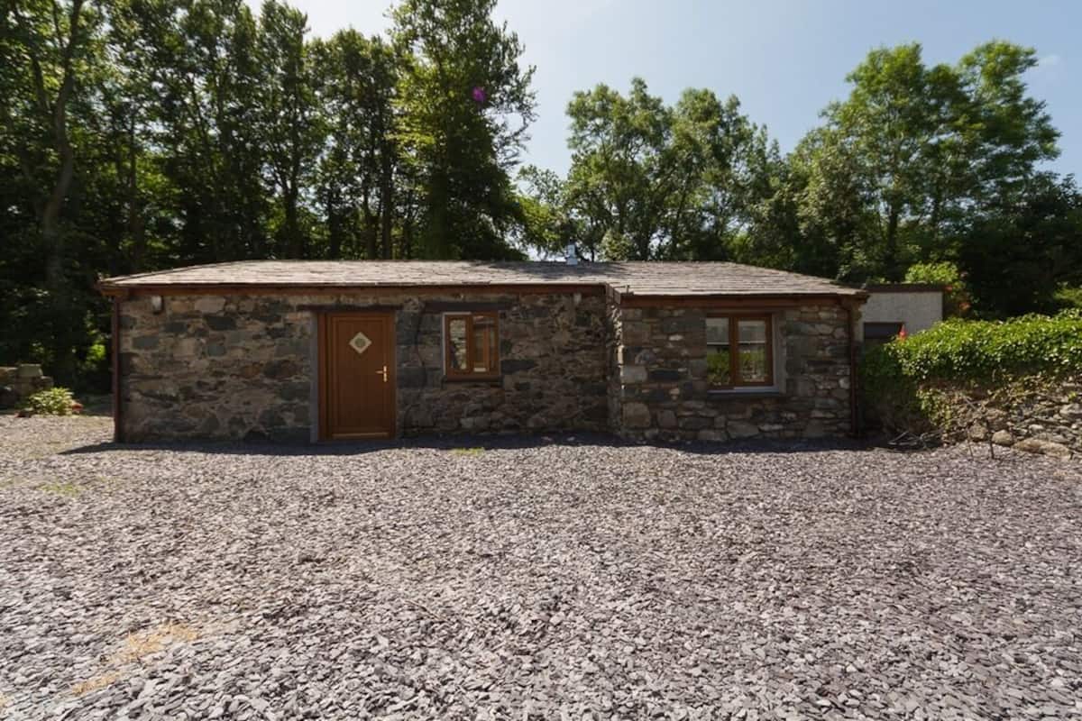 Une petite maison en pierre avec une porte et des fenêtres en bois, entourée d’une allée gravillonnée et d’arbres.