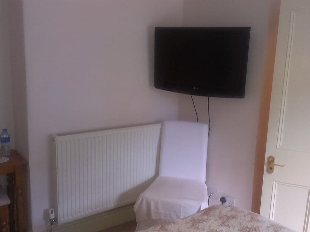 Une chambre à coucher avec un lit, un radiateur, une chaise blanche et un téléviseur fixé au mur.