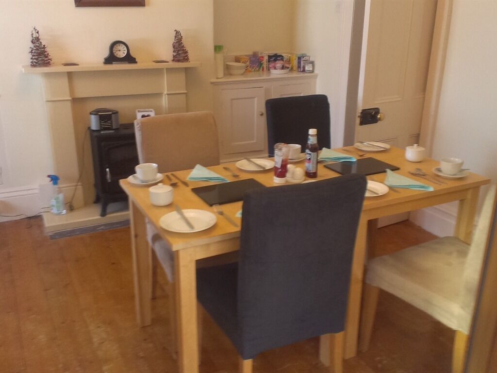 A dining area with a wooden table set for four, chairs, a fireplace, and a shelf with kitchen items.