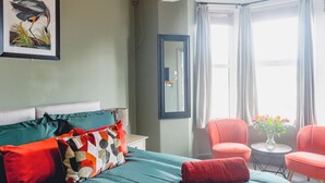 A hotel room with a bed, two chairs, a small table with flowers, and a large window with curtains. - Smarties Guest House (Newquay)