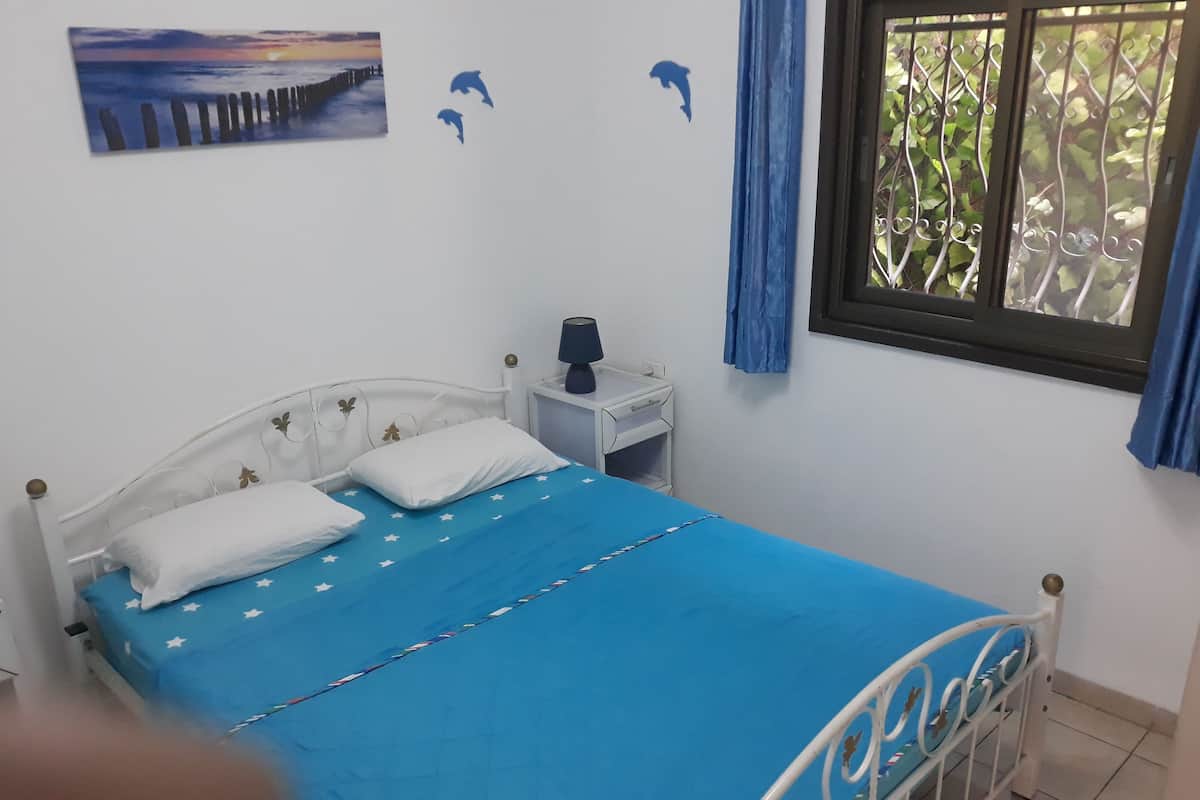 Un dormitorio con cama, mesita de noche, lámpara, ventana con cortinas y adornos en la pared, incluyendo una pintura de atardecer y siluetas de delfines.