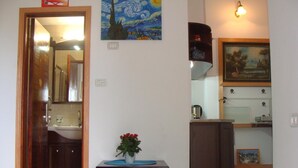 A room with a wooden door, a small table with a potted plant, a painting on the wall, and a kitchen area with a kettle. - Tamer guest house (Haifa)