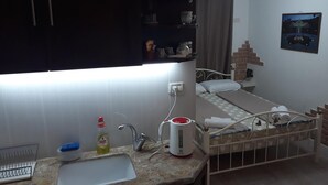 A small, well-lit room with a kitchenette area, a bed with a white headboard, and a wall-mounted picture. - Tamer guest house (Haifa)