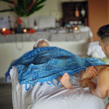 A person lying on a bed with a blue patterned blanket, another person sitting beside them, and a room with a visible bed, pillows, and a nightstand with items on it.