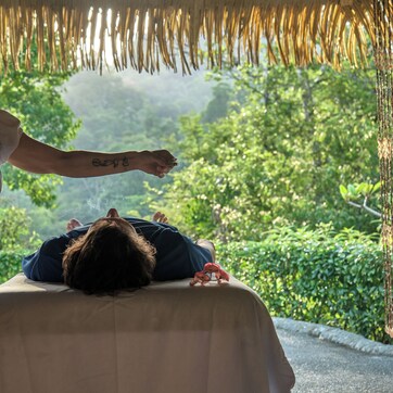 A person lying on a massage table outdoors, with a natural backdrop of trees and hanging decorations.