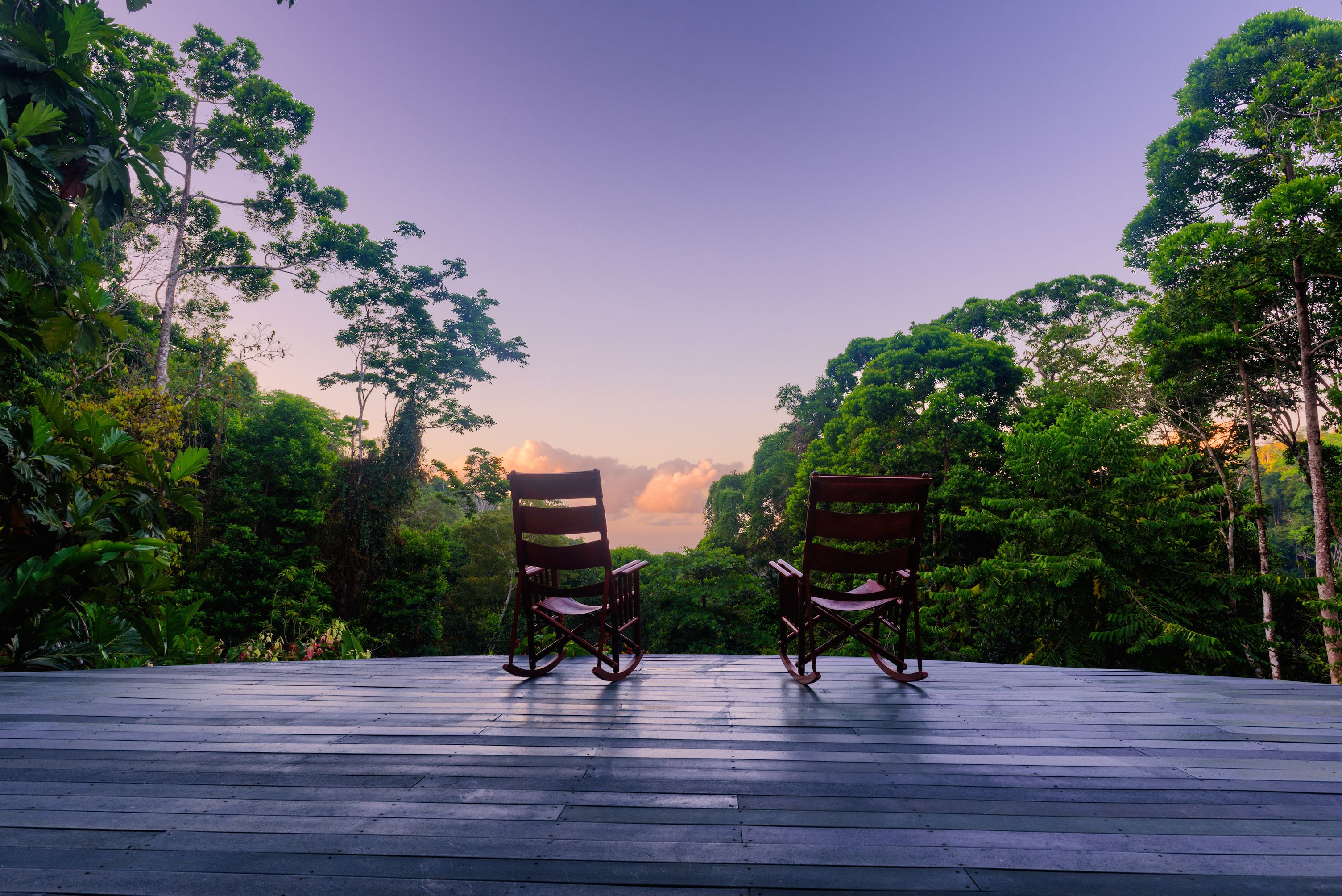 Dos mecedoras sobre una terraza de madera con vistas a una vegetación verde exuberante y un cielo despejado.