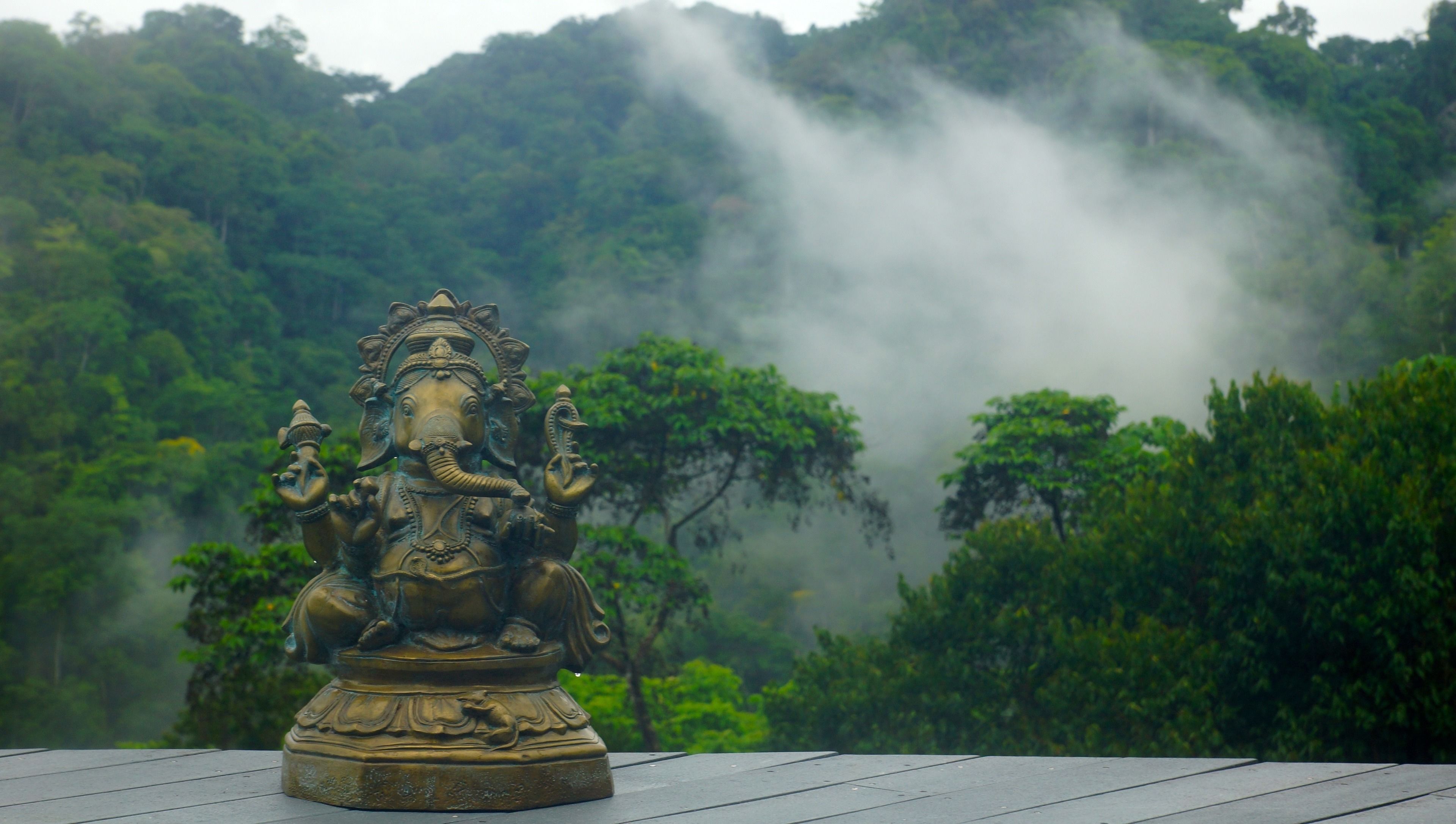 A statue of a deity on a wooden platform with a forested mountain background.