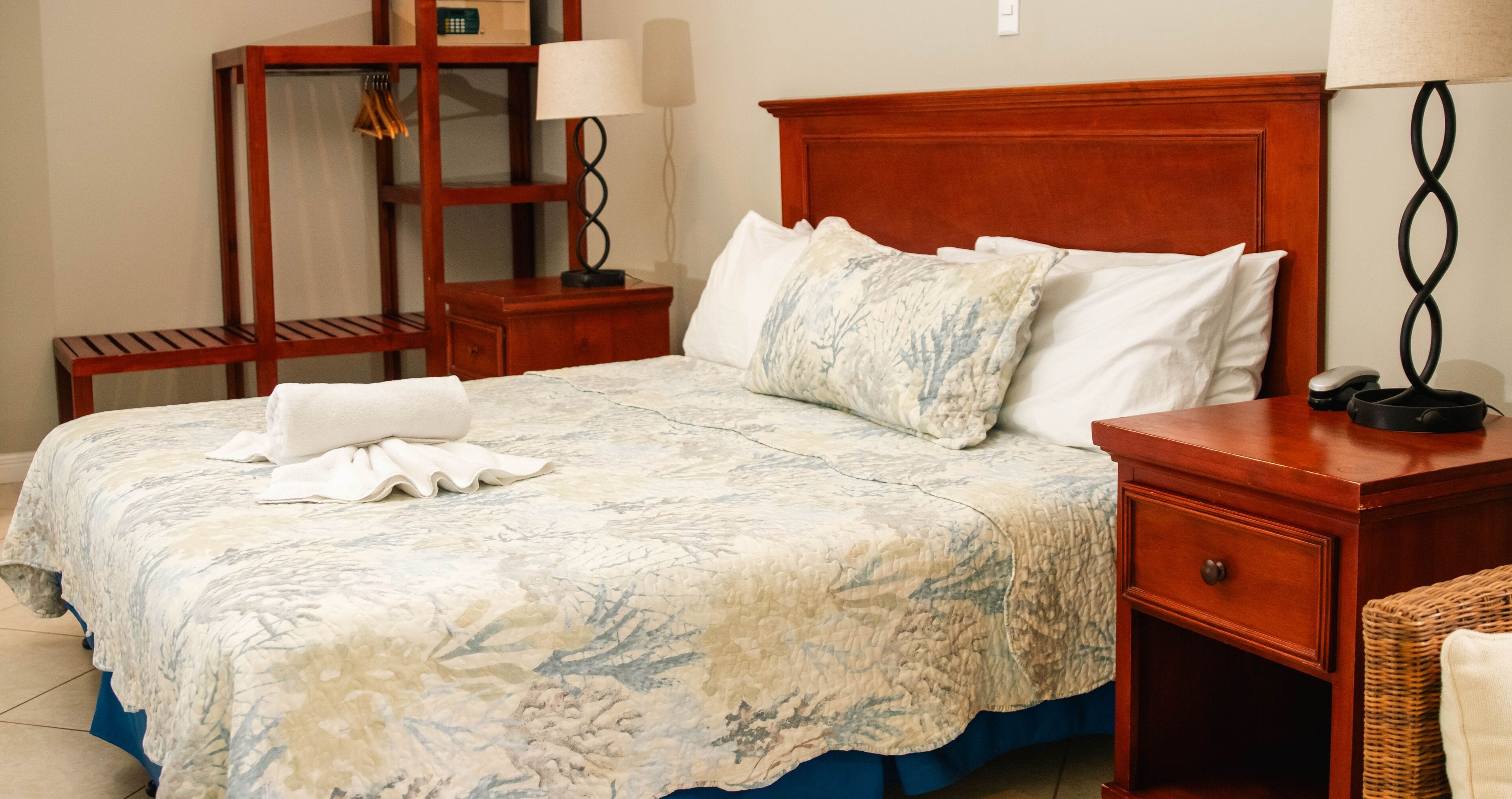 Una cama bien hecha con una colcha estampada, una mesita de noche de madera con una lámpara, y una silla de mimbre.