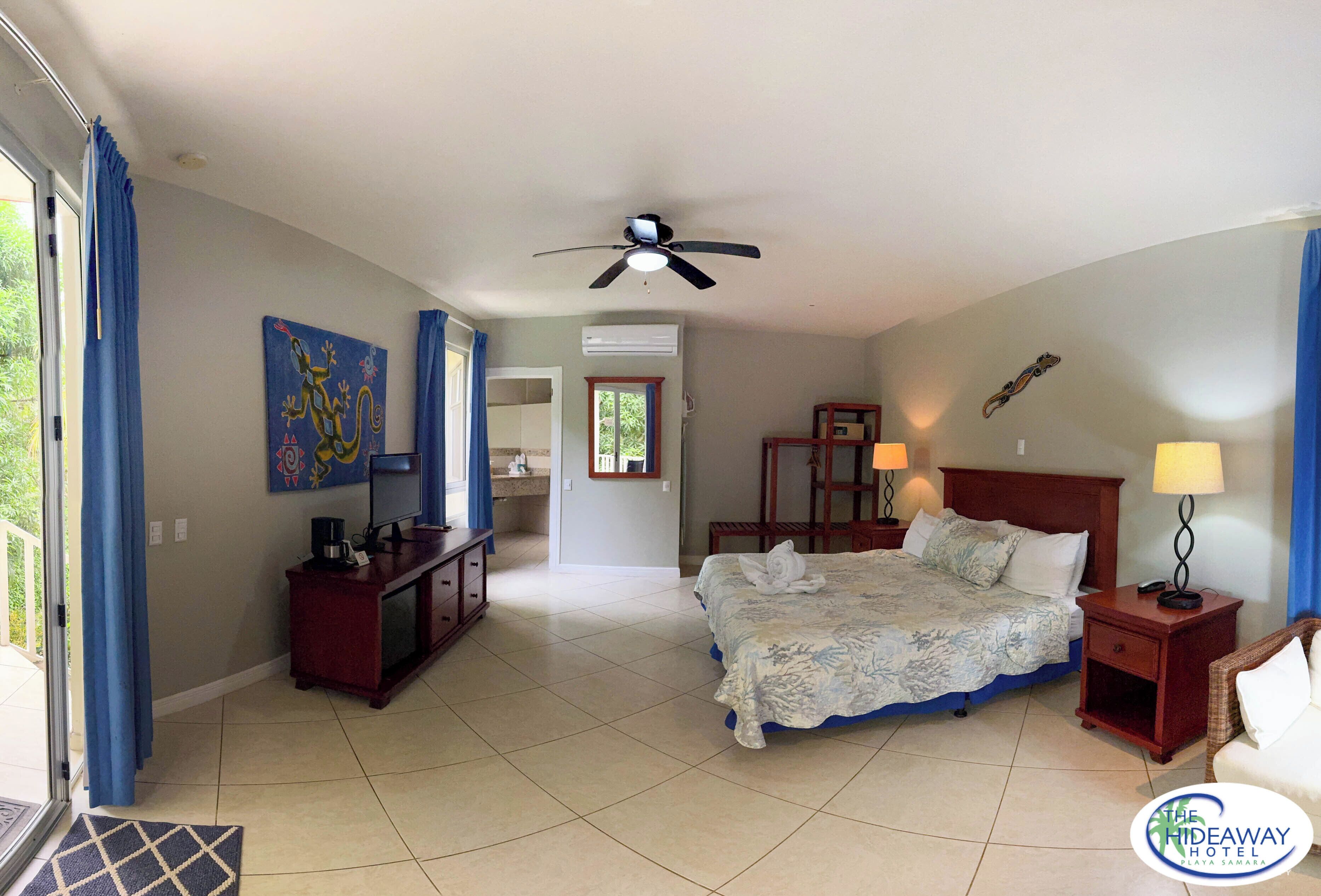 Una habitación de hotel con cama, escritorio, silla, mesita de noche, lámpara, ventilador de techo y obras de arte en las paredes.
