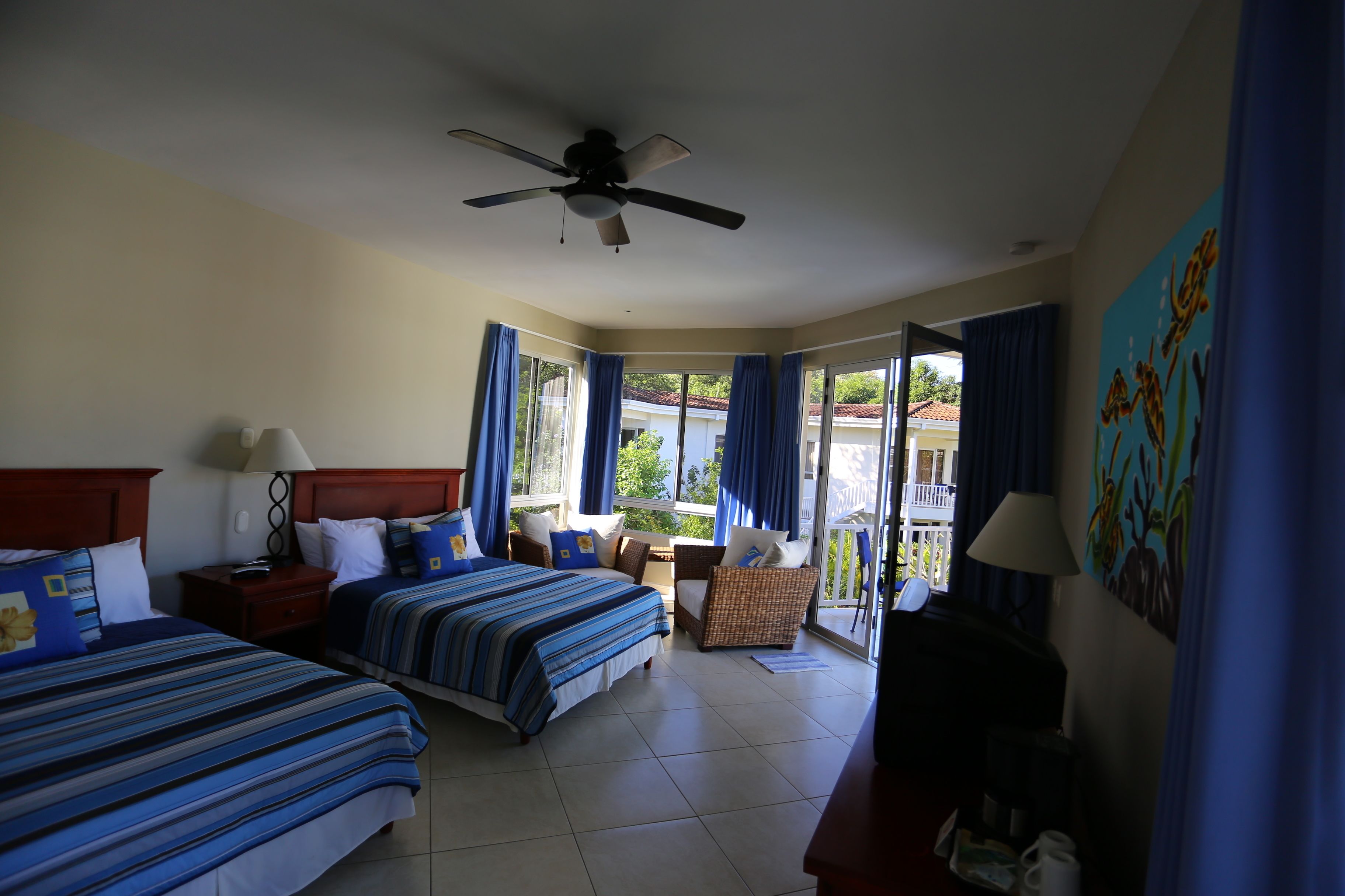 A hotel room with two beds, a ceiling fan, a balcony with blue curtains, and a painting on the wall.
