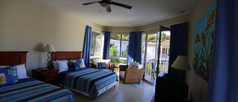 A hotel room with two beds, a ceiling fan, a balcony with blue curtains, and a painting on the wall.