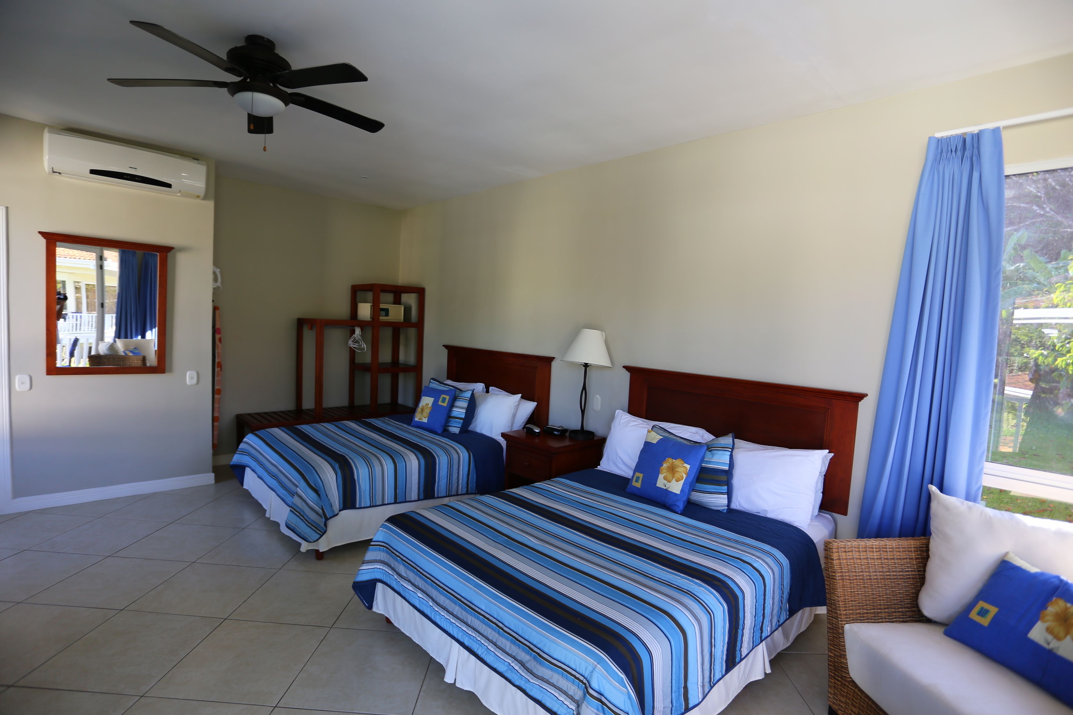 Habitació d'hotel amb dos llits, un ventilador de sostre, una finestra amb cortines i vistes a vegetació.