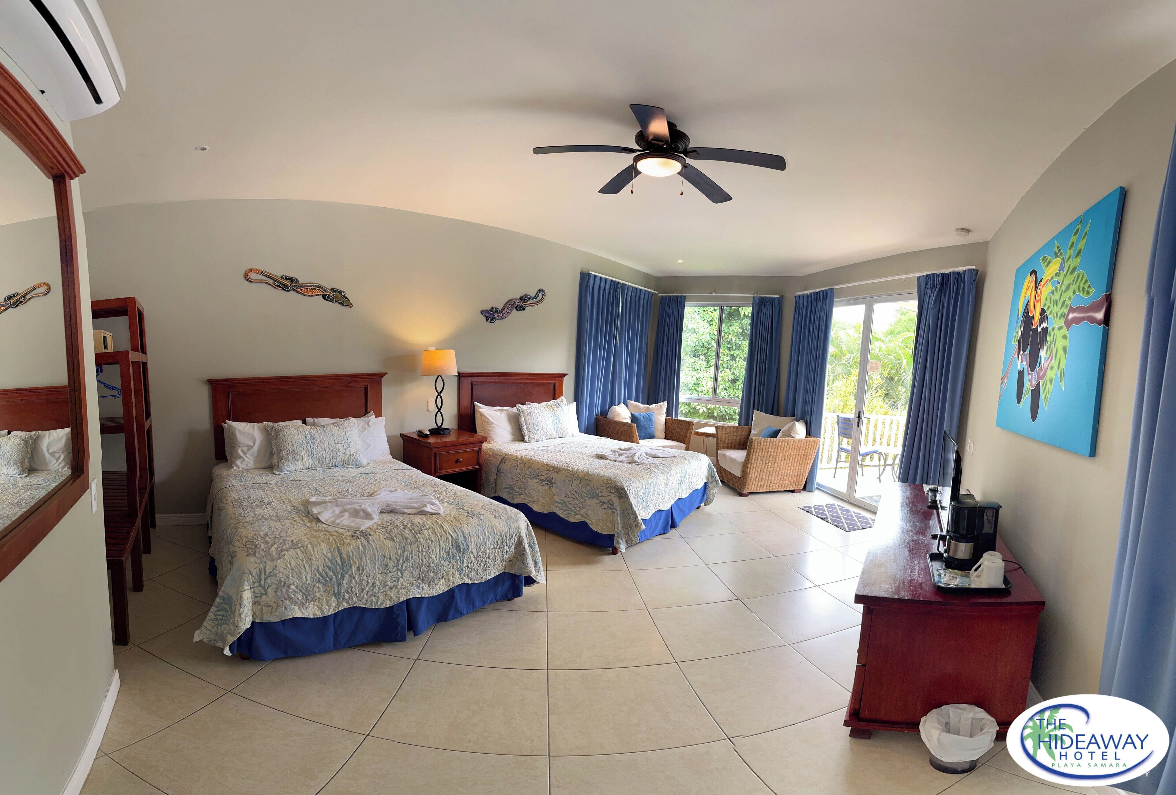 A hotel room with two beds, a desk, a chair, a ceiling fan, and a view of the outdoors.